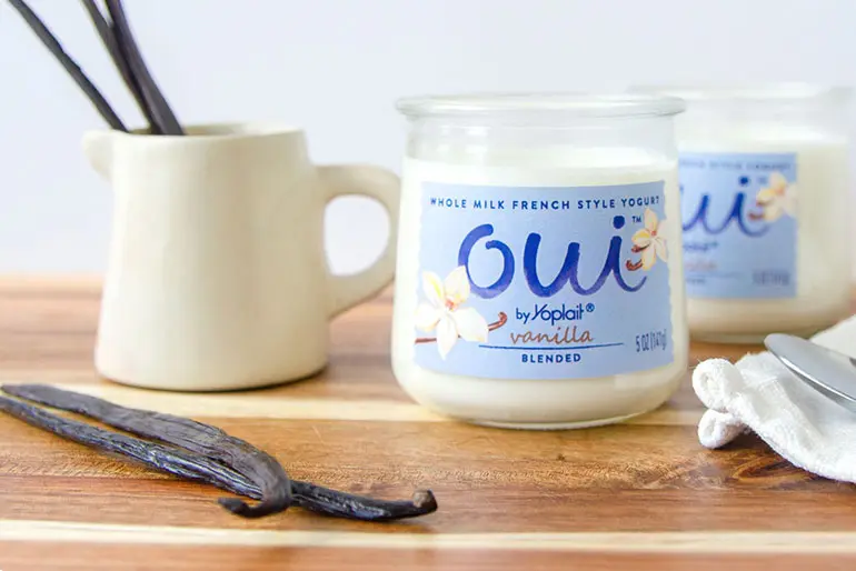 A jar of Vanilla Oui by Yoplait yogurt on a wood surface next to raw vanilla bean pods.