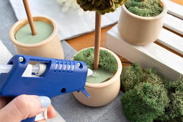Hand holding hot glue gun and gluing artificial moss on top of green foam with craft topiary in a Oui by Yoplait yogurt glass pot.