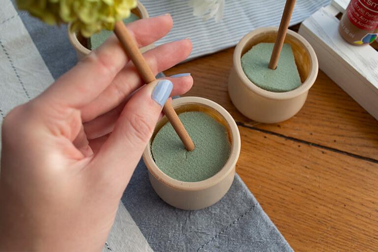 Hand placing a craft stick into a piece of green foam in a Oui by Yoplait yogurt painted jar.