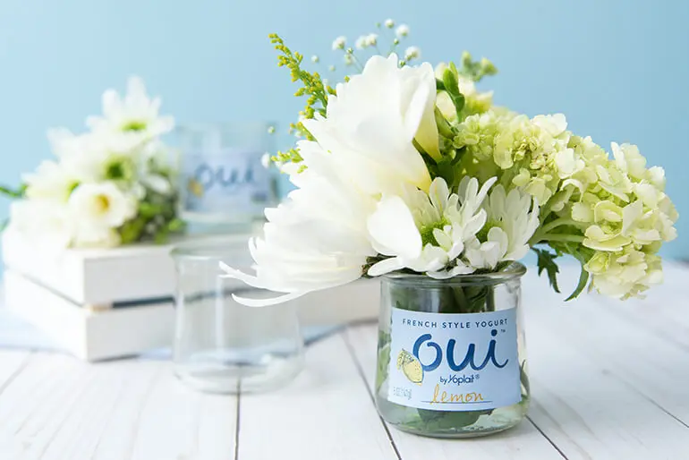 A Oui by Yoplait yogurt jar with flowers in it behind are more of the same flowers in a wooden crate.