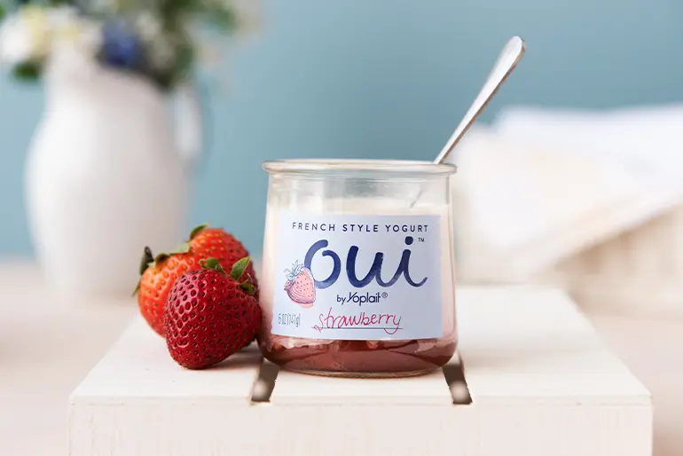 An open jar of strawberry Oui by Yoplait yogurt with spoon next to fresh strawberries.