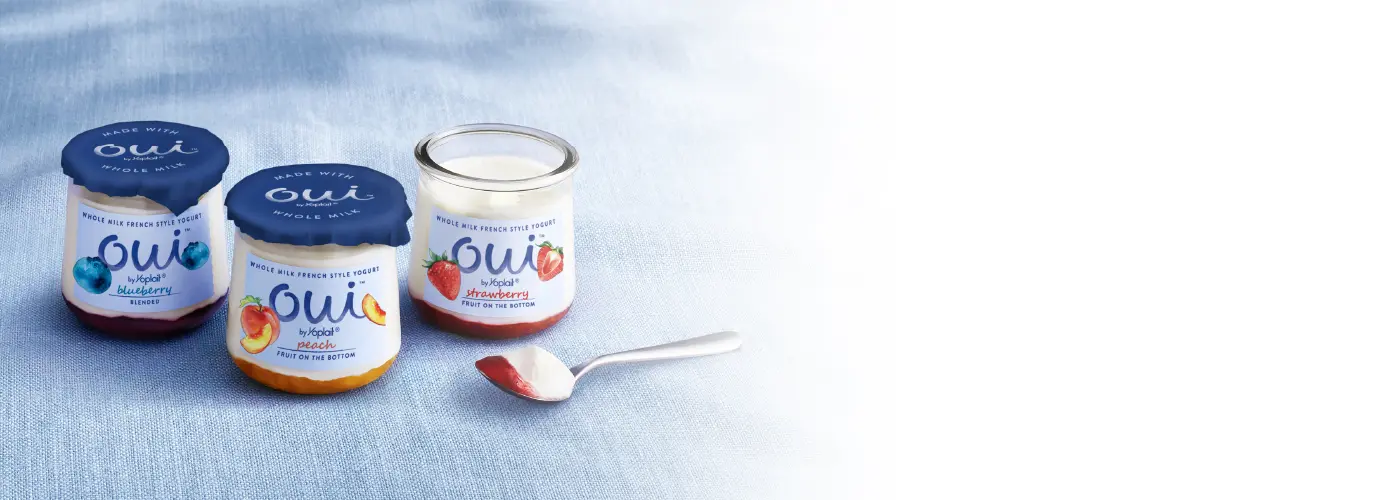 Jars of blueberry, peach and strawberry Oui by Yoplait yogurt on a blue tablecloth.