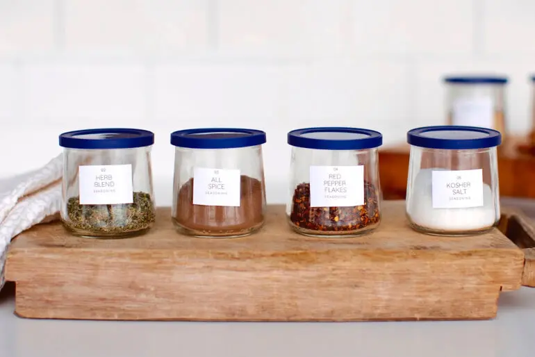 Three glass pots of Oui that have been upcycled to become spice jars with each labelled appropriately, sitting on a wooden chopping board.