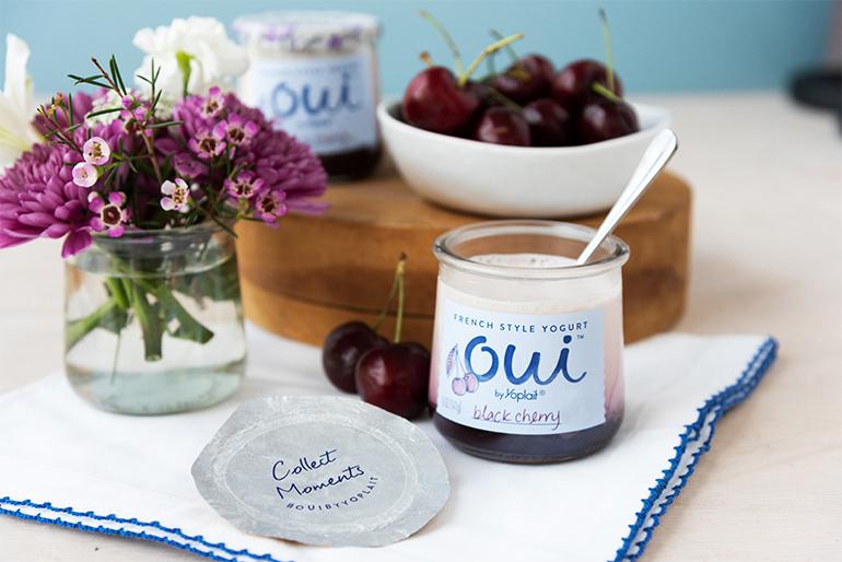A jar of Oui by Yoplait Black Cherry yogurt next to a bowl of black cherries and a jar of flowers.