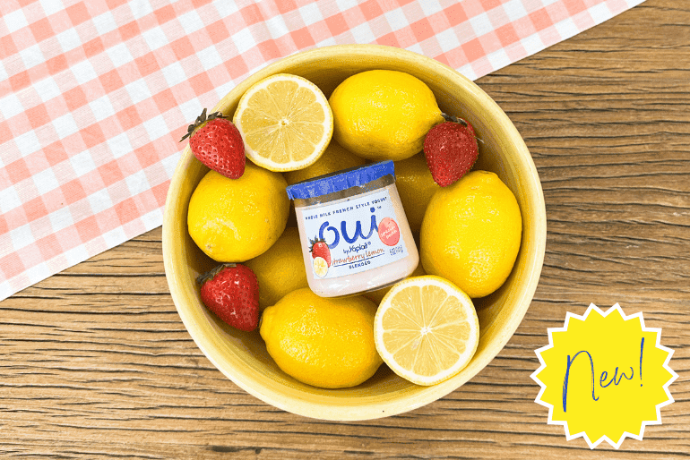 Overhead shot of a pale yellow bowl brimming with whole lemons, lemon halves, and fresh strawberries. Nestled amongst the fruit is a small jar of OUI by Yoplait Strawberry Lemon yogurt. The bowl rests on a rustic wooden surface partially covered by a pink and white gingham tablecloth. A bright yellow sunburst graphic with the word "New!" in dark blue script is positioned in the lower right corner.