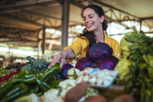 Smiling woman with dark hair wearing a yellow shirt reaching over a table full of colorful vegetables to grab a green pepper.